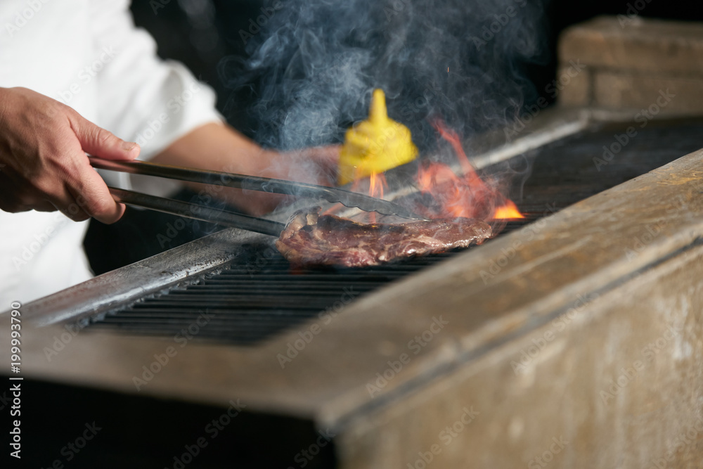 Young chef in a white apron standing near the brazier with coals. Man cooking beef steak on barbeque in the interior of modern professional kitchen Young chef in a white apron standing near the brazier with coals. Man cooking beef steak on barbeque in the interior of modern professional kitchen