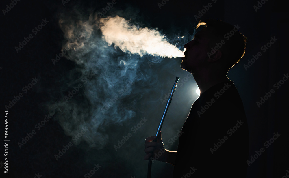Beautiful young man profile in backlight smoking hookah with a lot of smoke isolated black background. Club party menu concept