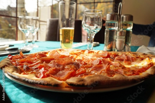 Whole ham pizza on a restaurant table, in the back there are glasses, beer, salt shaker and olive and vinegar bottles