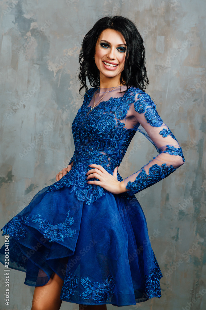 Φωτογραφία brunette woman in the blue evening dress is posing near the light texture wall background