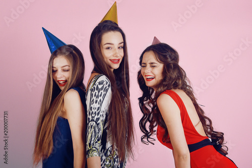 Crazy party time of three beautiful stylish women in elegant evening outfit celebrating , having fun, dancing smile on white background. Best friends girls in party cap on pink background.