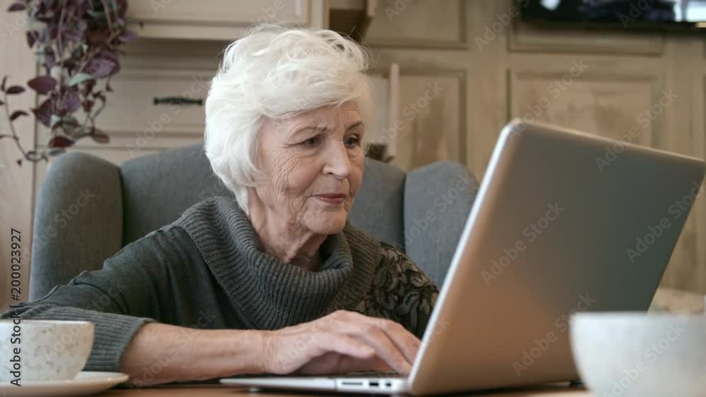 Beautiful senior lady sitting at table in restaurant and typing on modern laptop Beautiful senior lady sitting at table in restaurant and typing on modern laptop
