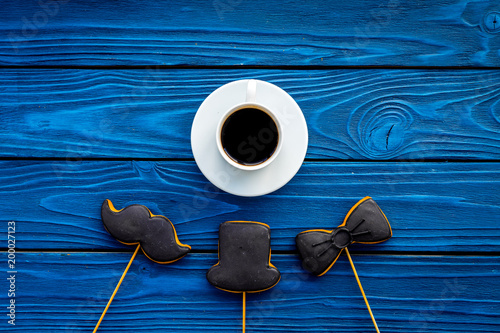 Men's birthday celebration concept. Cookies in shape of moustache, hat, bow tie near coffee on blue wooden background top view copy space