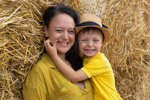 Happy family. A young woman with dark hair hugs her son in a hat against a background of hay. Mom and son. © AnastazjaSoroka