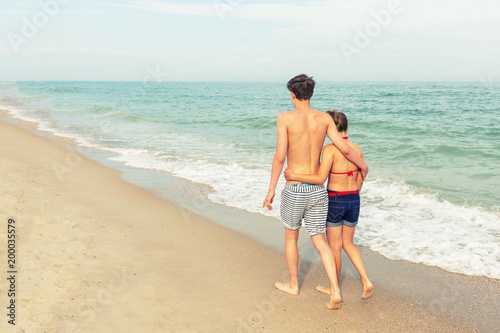 Two teenagers: a girl and a boy with blond hair, dressed in a swimsuit are walking on a sea beach. Back view.