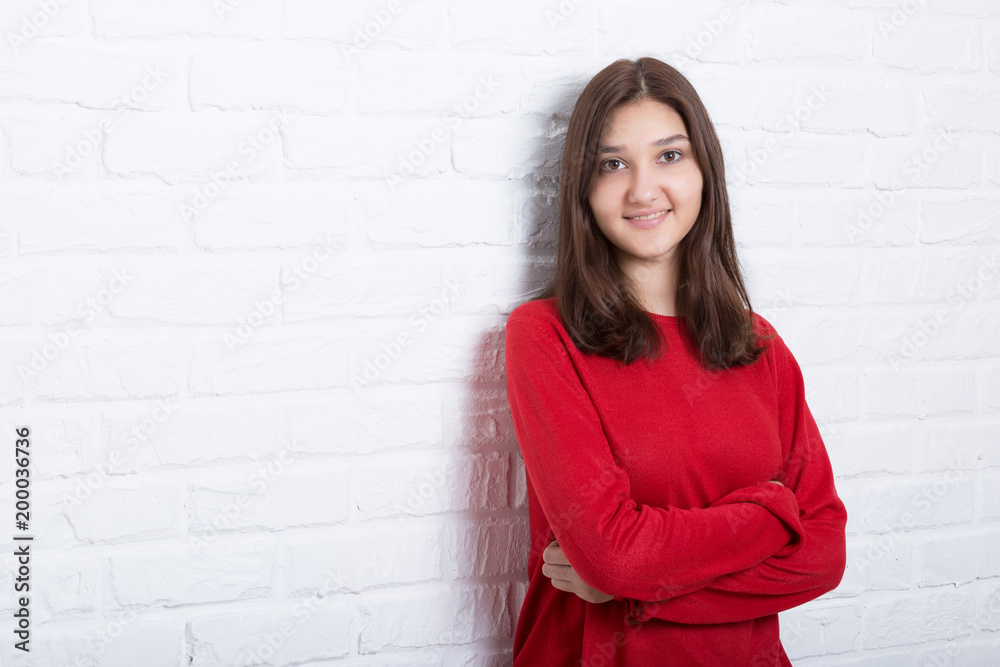 A young pretty smiling girl in a red sweater and a hat against a modern white brick wall in the loft style. She looks at the viewer. Copy space. A young pretty smiling girl in a red sweater and a hat against a modern white brick wall in the loft style. She looks at the viewer. Copy space.