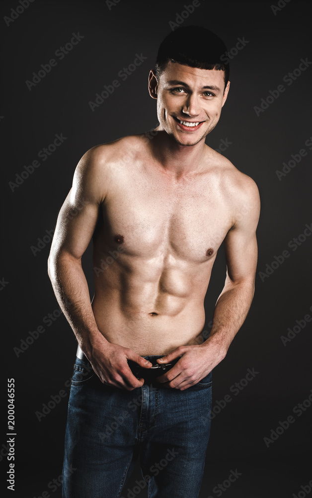 Fototapeta premium Portrait of cheerful young man with attractive body holding hands on belt. Happy bare-chested guy concept