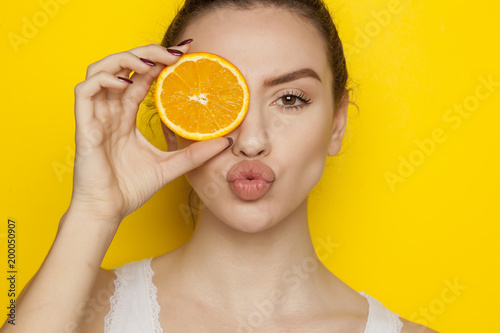 Young woman posing with slice of orange on her face on yellow background