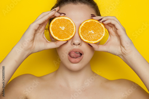 Young sexy woman posing with slices of oranges on her face on yellow background