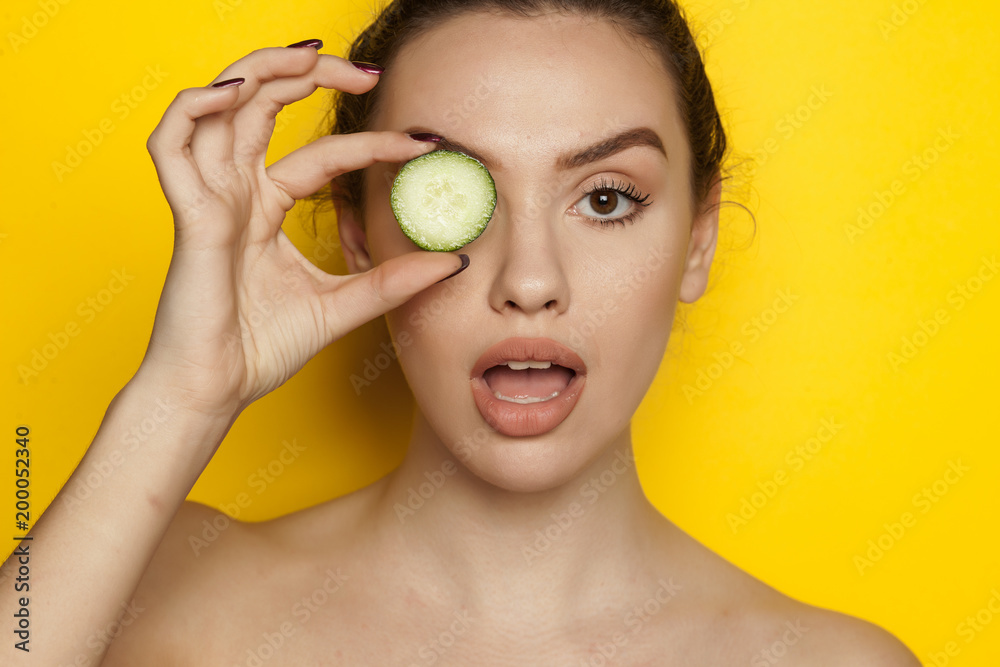 Surprised young woman posing with slice of cucumber on her face on yellow background Surprised young woman posing with slice of cucumber on her face on yellow background