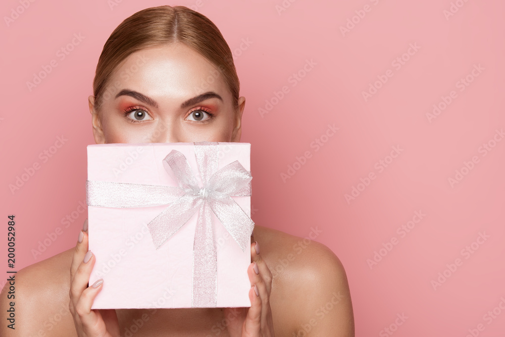 Beautiful woman with eye makeup and nude shoulders covering lower face with gift box. Copy space in right side. Isolated on rose background Beautiful woman with eye makeup and nude shoulders covering lower face with gift box. Copy space in right side. Isolated on rose background