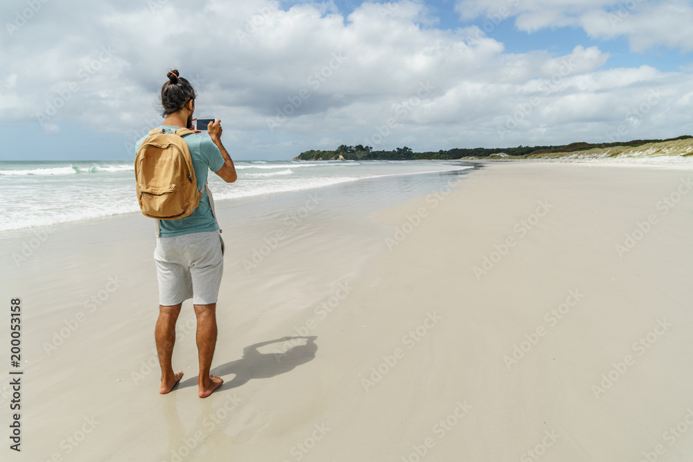 rear view of man taking photo of coastline with smartphone, Rarawa beach, New Zealand