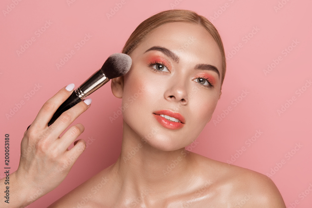 Portrait of peaceful attractive woman with maquillage and combed hair. Female hand with blush brush is in frame. Isolated on rose background Portrait of peaceful attractive woman with maquillage and combed hair. Female hand with blush brush is in frame. Isolated on rose background