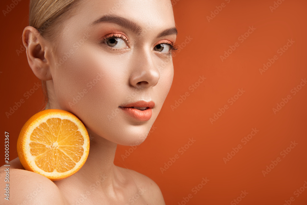 Close up portrait of young beauty having healthy skin. She is holding half of fresh orange on her shoulder and looking at camera with tranquility. Copy space in right side. Isolated on background Close up portrait of young beauty having healthy skin. She is holding half of fresh orange on her shoulder and looking at camera with tranquility. Copy space in right side. Isolated on background