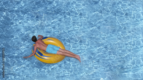 Young man is floating on yellow inflatable air ring/circle in pool with blue water, vacation concept with space for text. Top view.