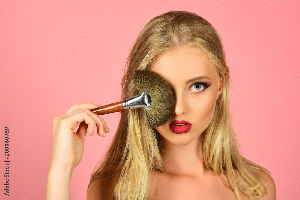 Obraz premium Seductive girl with clean fresh skin, red lips, blonde hair covers eye with makeup brush. Skincare, cosmetology, beauty, fashion - sexy blonde model with healthy skin, beautiful makeup. Copy space.