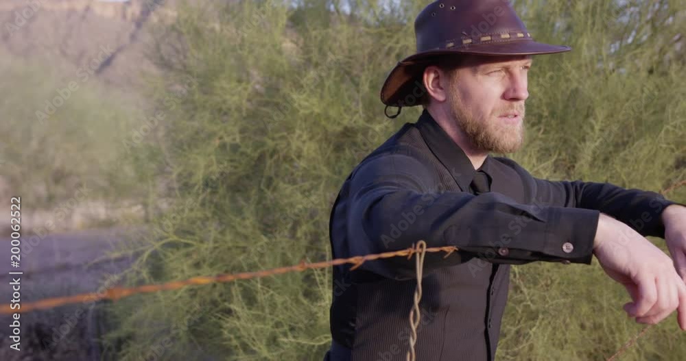 American cowboy leans on barbwire fence looking out towards heard in sunset?