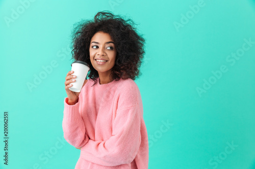 Portrait of american woman 20s with afro hairdo looking aside while drinking takeaway coffee or tea from paper cup, isolated over blue background