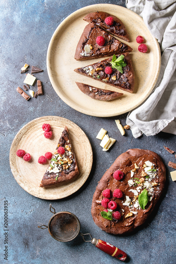 Whole and slised homemade dessert chocolate pizza with different chocolates, raspberries and mint served on ceramic pale with cloth and ingredients above over blue texture background. Top view, space.