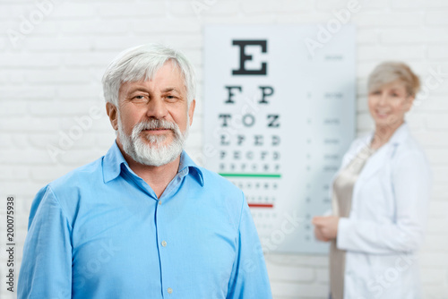 Old patient staying in front of ophthalmologist. Looks satisfied. wearing blue shirt. trying to improve health and vision. Doctor behind looking friendly and expirienced.