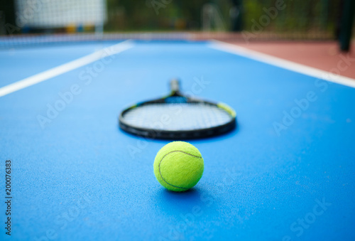 Frontview of yellow tennis ball is laying near professional racket on blue carpet of opened tennis cort. Contrast image with satureted colors and shadows. Concept of sport equipment photo. photo