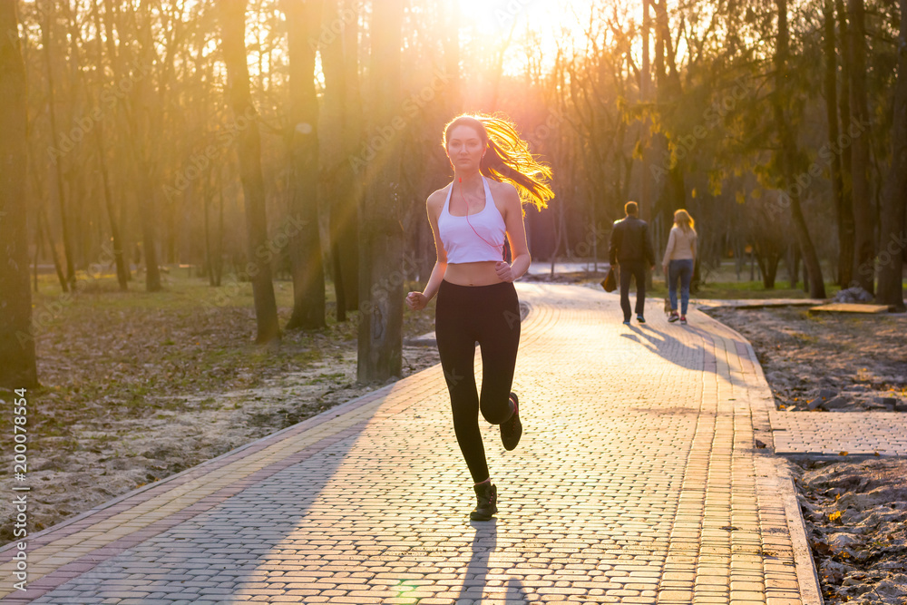 Obraz premium Young girl in headphones enjoys jogging in park at sunset