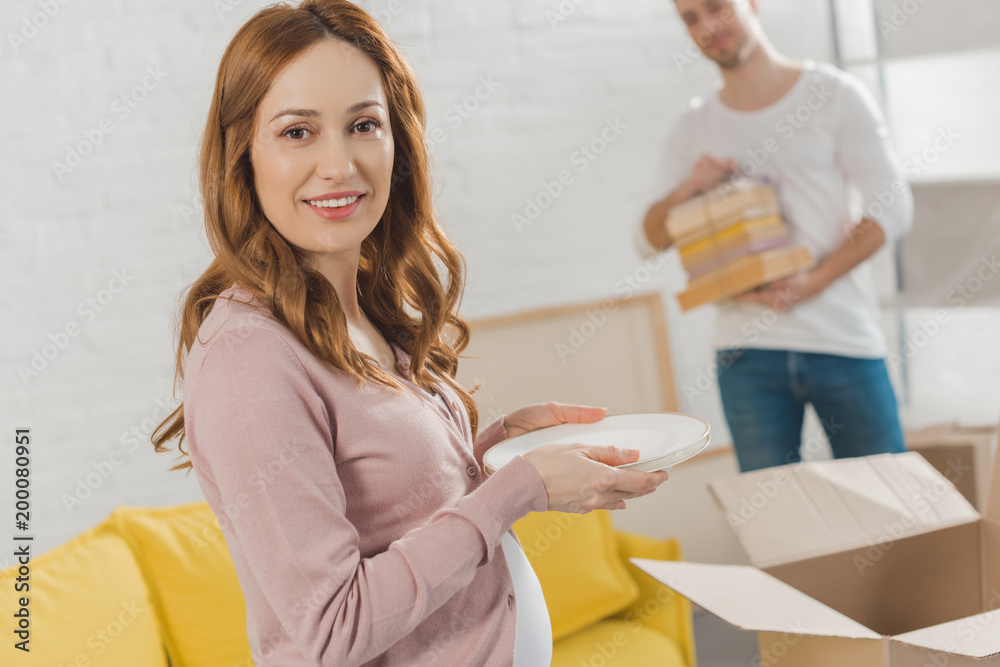 happy pregnant woman holding dishware and smiling at camera while relocating with husband happy pregnant woman holding dishware and smiling at camera while relocating with husband