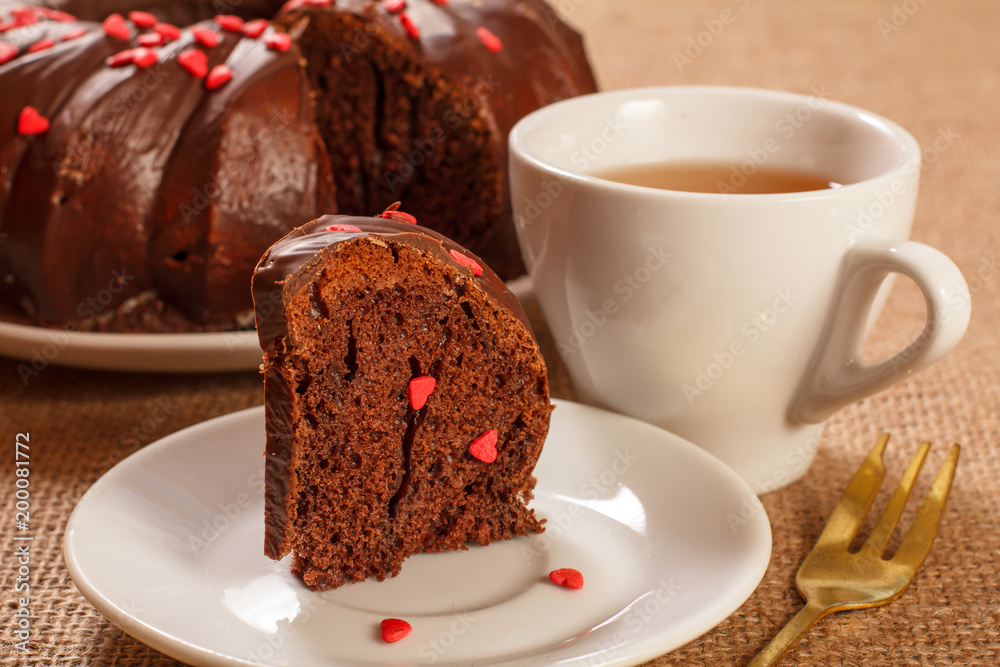 Slice of delicious homemade chocolate cake decorated with small hearts made of caramel on saucer and cup of tea on table with sackcloth Slice of delicious homemade chocolate cake decorated with small hearts made of caramel on saucer and cup of tea on table with sackcloth