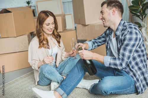 happy young couple celebrating relocation with champagne while sitting near cardboard boxes in new home