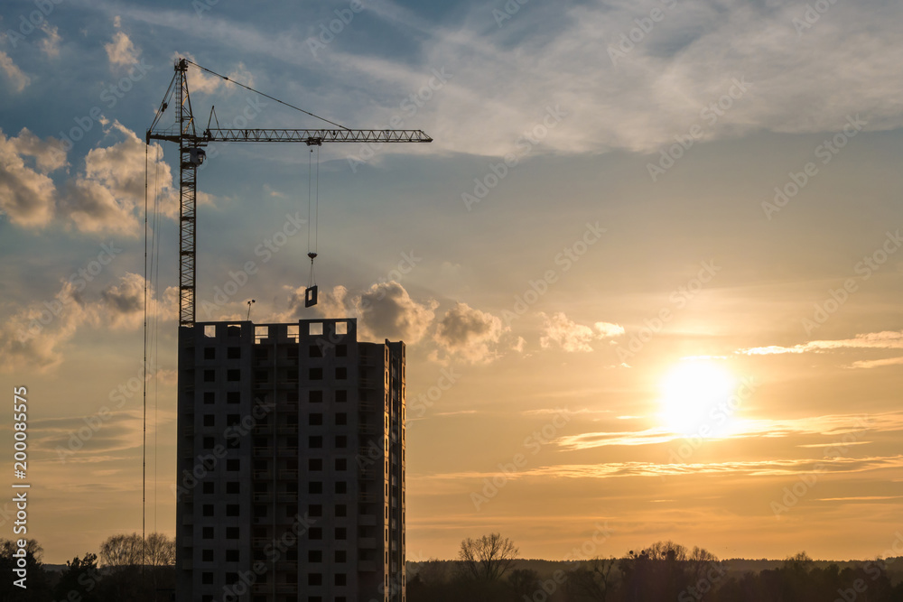 Tower cranes and unfinished multi-storey high near buildings under construction site in the rays of the setting sun of beautiful sunset Tower cranes and unfinished multi-storey high near buildings under construction site in the rays of the setting sun of beautiful sunset