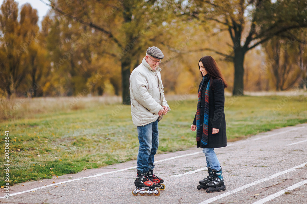 Fototapeta premium A happy pensioner stands on roller skates on the road with his daughter and enjoys good weather in the autumn park.