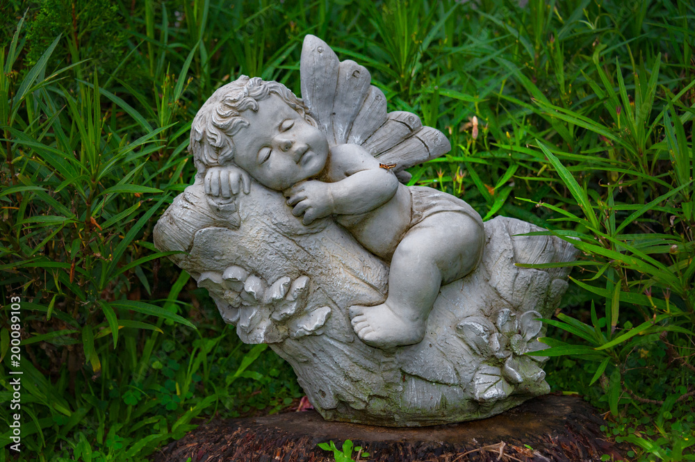 LITTLE ANGEL SLEEPS. Sculpture of a little angel sleeping on tree a branch. It is placed on top of an old tree stump surrounded with green leaves. LITTLE ANGEL SLEEPS. Sculpture of a little angel sleeping on tree a branch. It is placed on top of an old tree stump surrounded with green leaves.