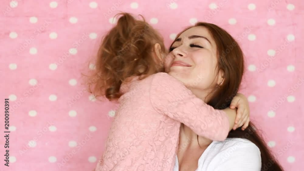 Mother and daughter embrace, smiles, have fun, laugh and kissed. Close-up portrait young woman and her child on pink background.
