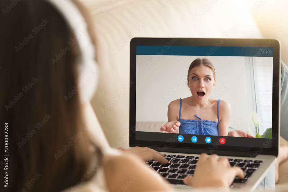 Two girlfriends chatting on laptop via video call application. Girl on  screen has surprised expression. Friend tells private secret, chat via  webcam, virtual communication. Close up, focus on screen. Stock Photo
