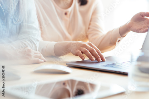 Close up shot of two female businesswomen working with hands on a laptop keyboard in an office in sunlight at a wooden table