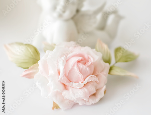 Realistic Fabric Silk flower in light pink cream and white colors rose hand made on white blurred background with angel or amur statuette. Vintage retro style card or decoration