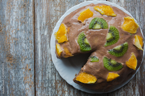 Homemade sponge cake with chocolate cacao ganache frosting. Slices of kiwi and orange on the dessert. Baked pastry in the kitchen on the old vintage wooden background.