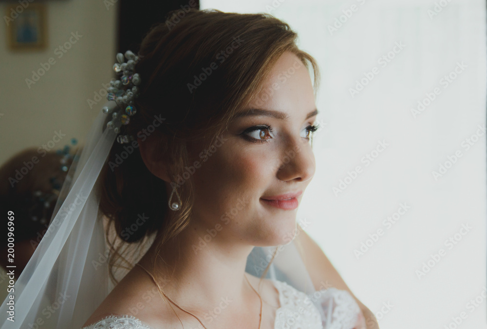Beautiful bride is waiting groom near the window. Young blonde girl in white wedding dress and elegant hair accessories. Wedding portrait with sun shades on face.