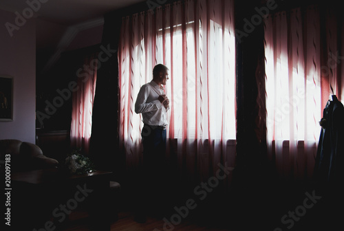 Handsome blond groom is standing near the window. Dark red curtains and sun lights on background. White wedding shirt and cufflinks. Groom waiting for his bride.