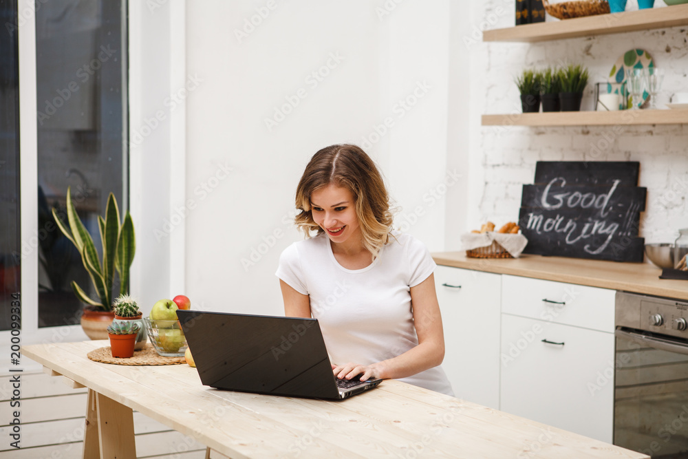 A beautiful girl is sitting at the table in the kitchen and looks at the laptop computer monitor, typing text. Modern technological devices. the Internet A beautiful girl is sitting at the table in the kitchen and looks at the laptop computer monitor, typing text. Modern technological devices. the Internet