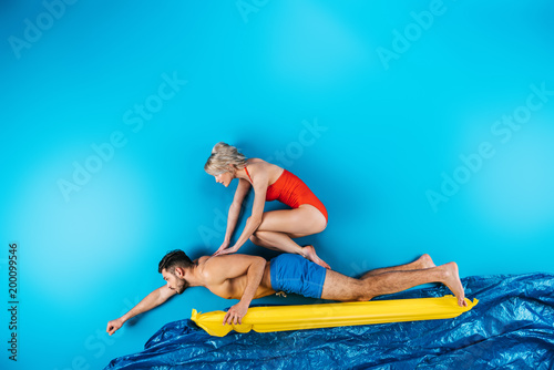 girl in swimsuit sitting on back of man swimming on inflatable mattress on blue