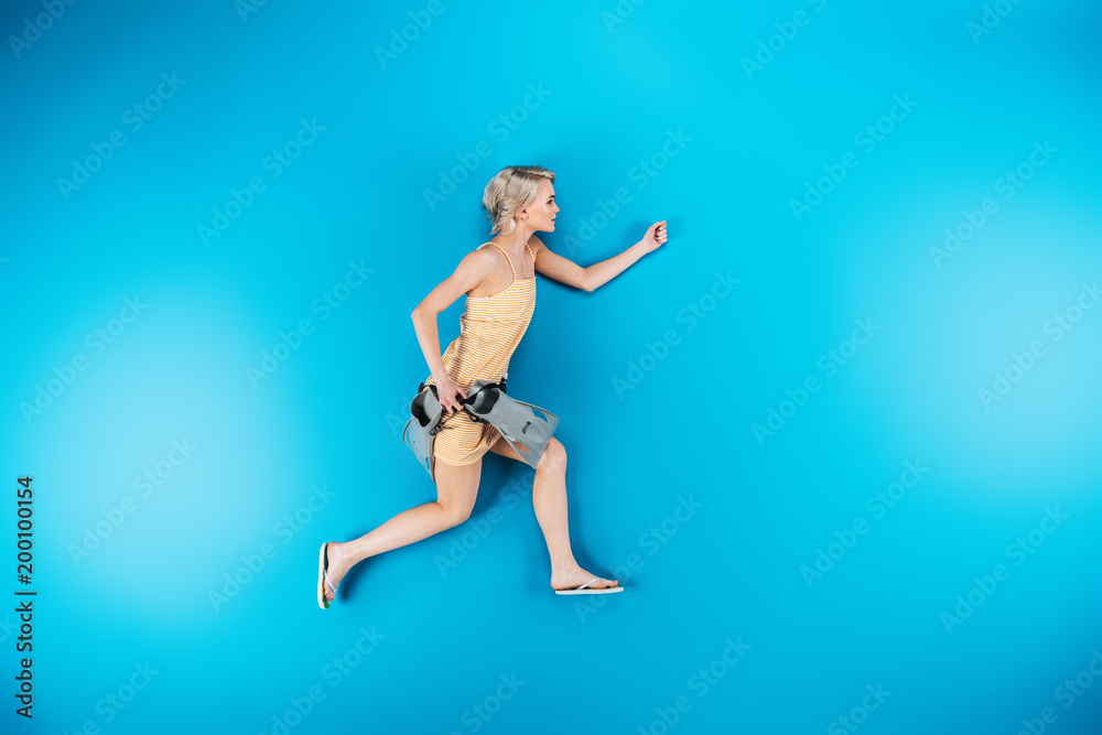 full length view of beautiful young woman in flip flops holding flippers and looking away on blue