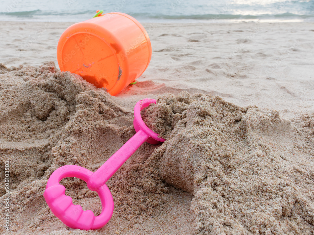 Pink spade and orange plastic bucket of sand tool toy set for kid and toddler on the sand over beach, sea, and sky background Pink spade and orange plastic bucket of sand tool toy set for kid and toddler on the sand over beach, sea, and sky background