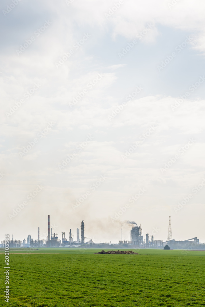 View of an oil refinery with smoking chimneys releasing toxic gases in the air, surrounded by crop fields in the french countryside under a pale sunlight.