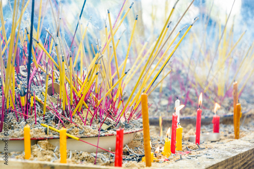 Close-up view of burning incense sticks planted among coins into a pile of sand on a table, diffusing a bluish smoke during a buddhist ceremony, with lighted candles in the foreground. © olrat