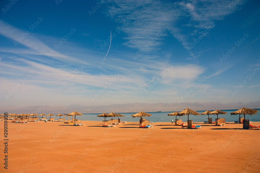 straw umbrellas from the sun on a beach on the shore of the red sea against a blue sky with white clouds and in the distance mountains in a haze on a sunny day