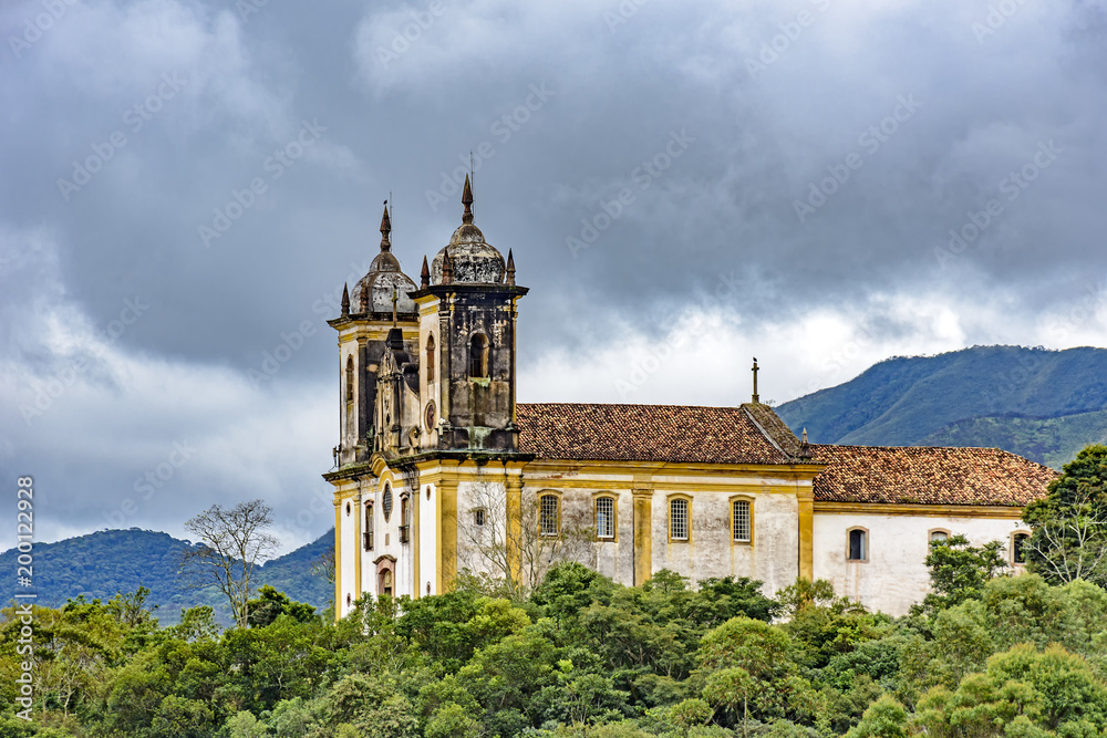Ancient historical church high in one of the several mountains of the city of Ouro Preto with the vegetation and dark clouds in background