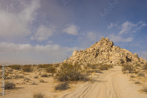 Soft sand road leads through desert to a massive hill made of sandstone boulders.