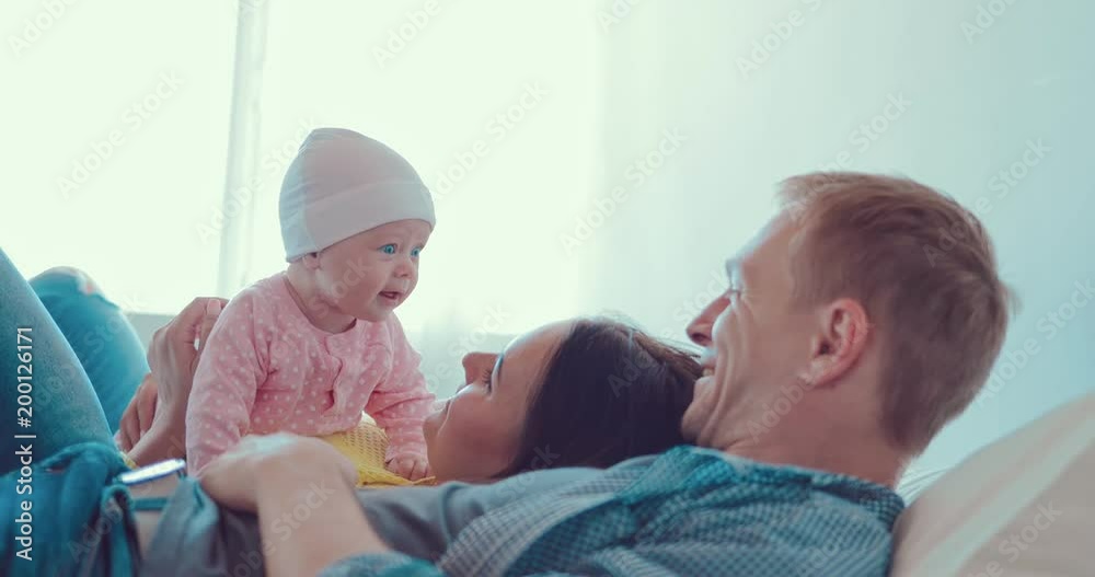Young and happy family is lying on the bed. Kid lies on his stomach and parents, looks into their eyes and smiles back Young and happy family is lying on the bed. Kid lies on his stomach and parents, looks into their eyes and smiles back