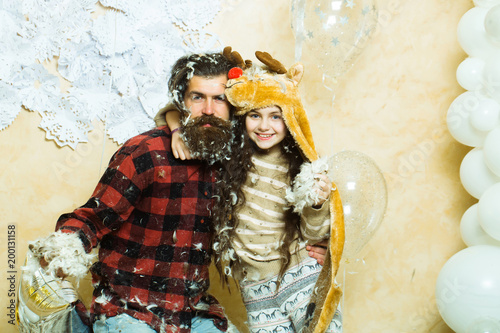 Happy christmas cute small girl or pretty child in winter hat and bearded man in checkered shirt with feathers on face, near silver and white balloons and decorative new year snowflakes